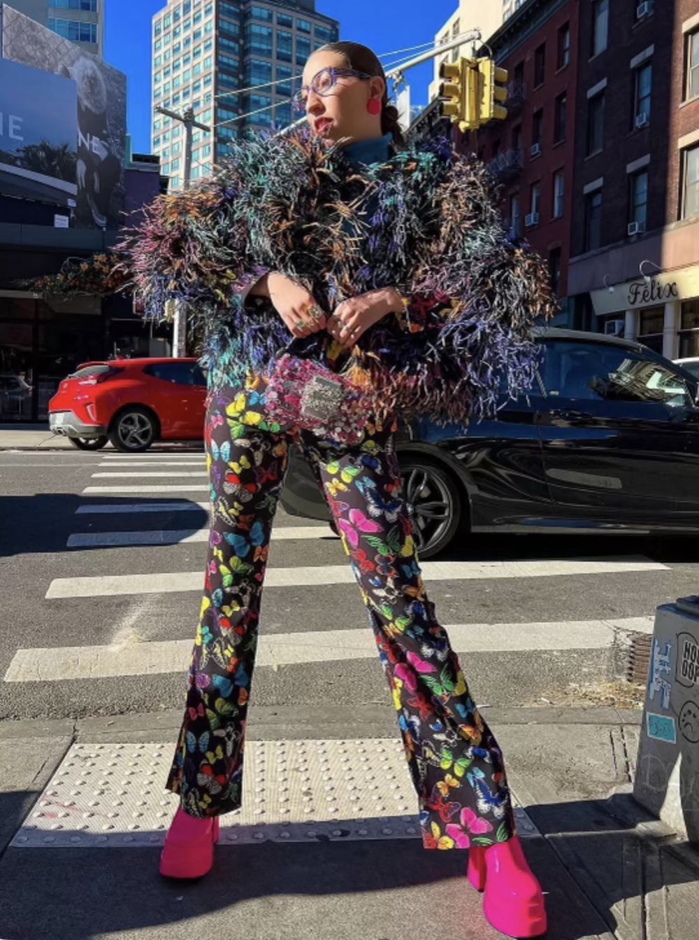 A woman in a multicolor feather jacket and butterfly print pants at a city crosswalk