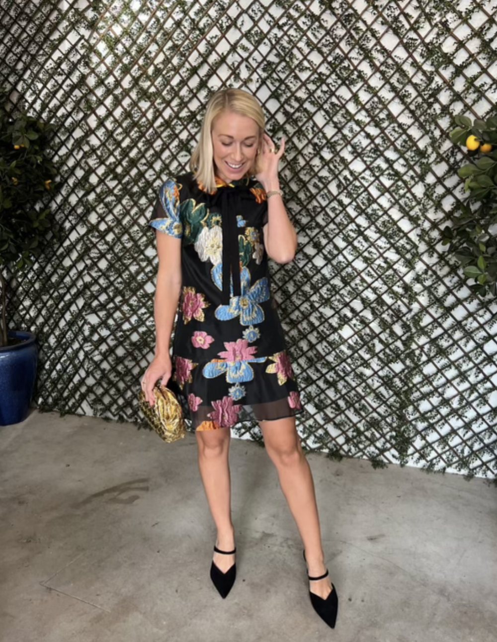 A woman in a black organza dress with metallic florals in front of a lattice wall