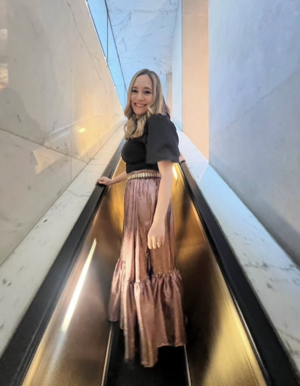 Lexie in a black top and pink metallic tiered skirt on an escalator